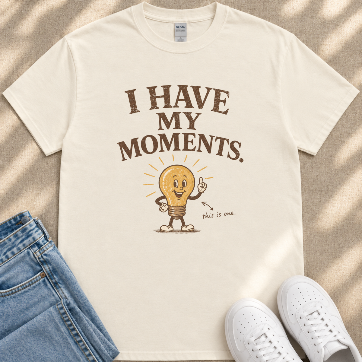 A white t-shirt with a lightbulb graphic and text, along with a pair of jeans and white sneakers, laid out on a beige surface.