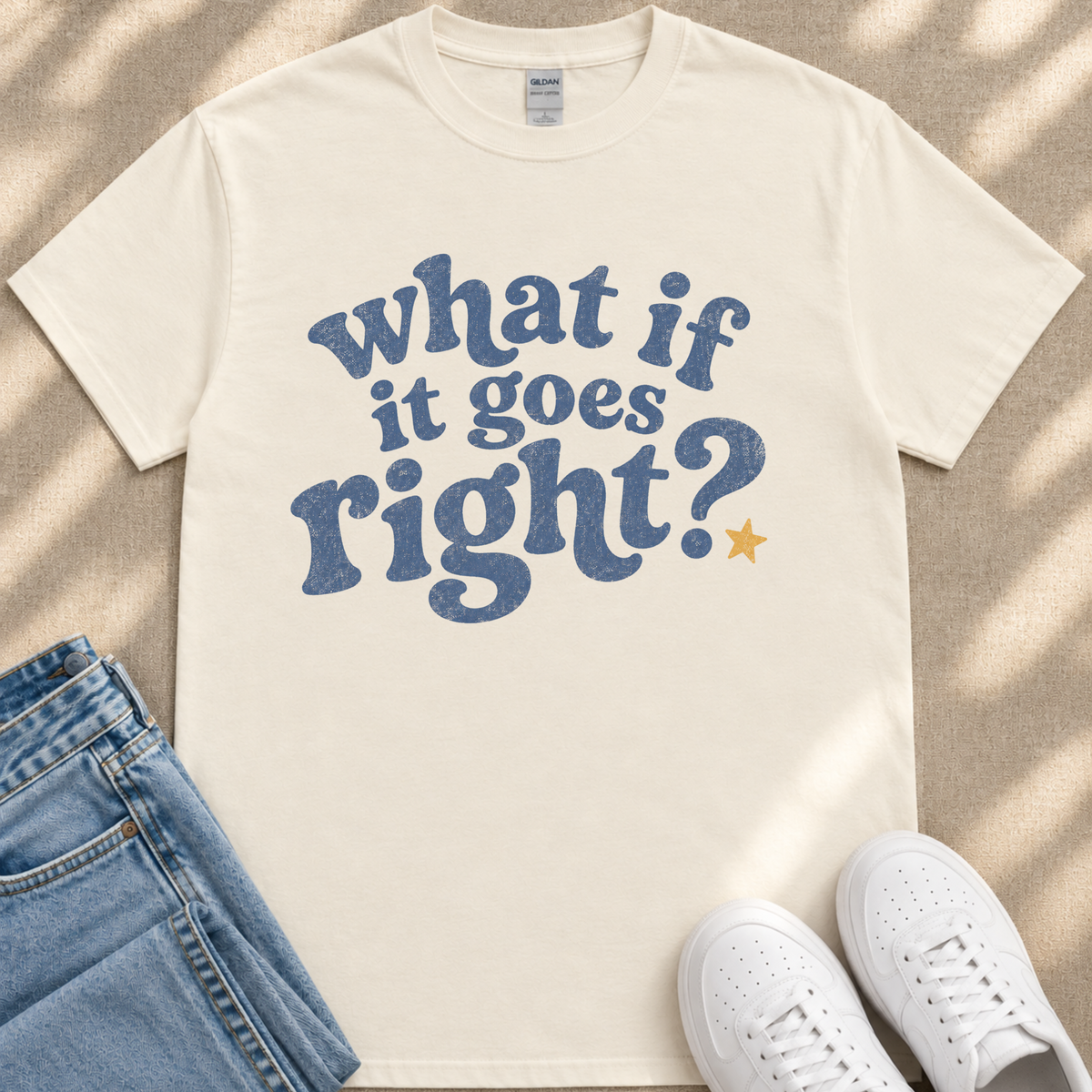 A white t-shirt with a blue text design that reads "What if it goes right?" along with a yellow star, placed on a beige surface next to a pair of blue jeans and white sneakers.
