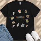 A black t-shirt with a colorful graphic design featuring various whimsical and nature-inspired icons, such as a mushroom, pine cone, butterfly, and more, along with the text "COLLECTING LITTLE JOYS". The t-shirt is placed on a beige surface, and there are a pair of jeans and white sneakers visible in the image.