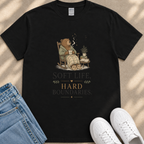 A black t-shirt with a graphic design depicting a bear sitting in a rocking chair, holding a cup and surrounded by various objects. The text below the image reads "Soft life, hard boundaries."