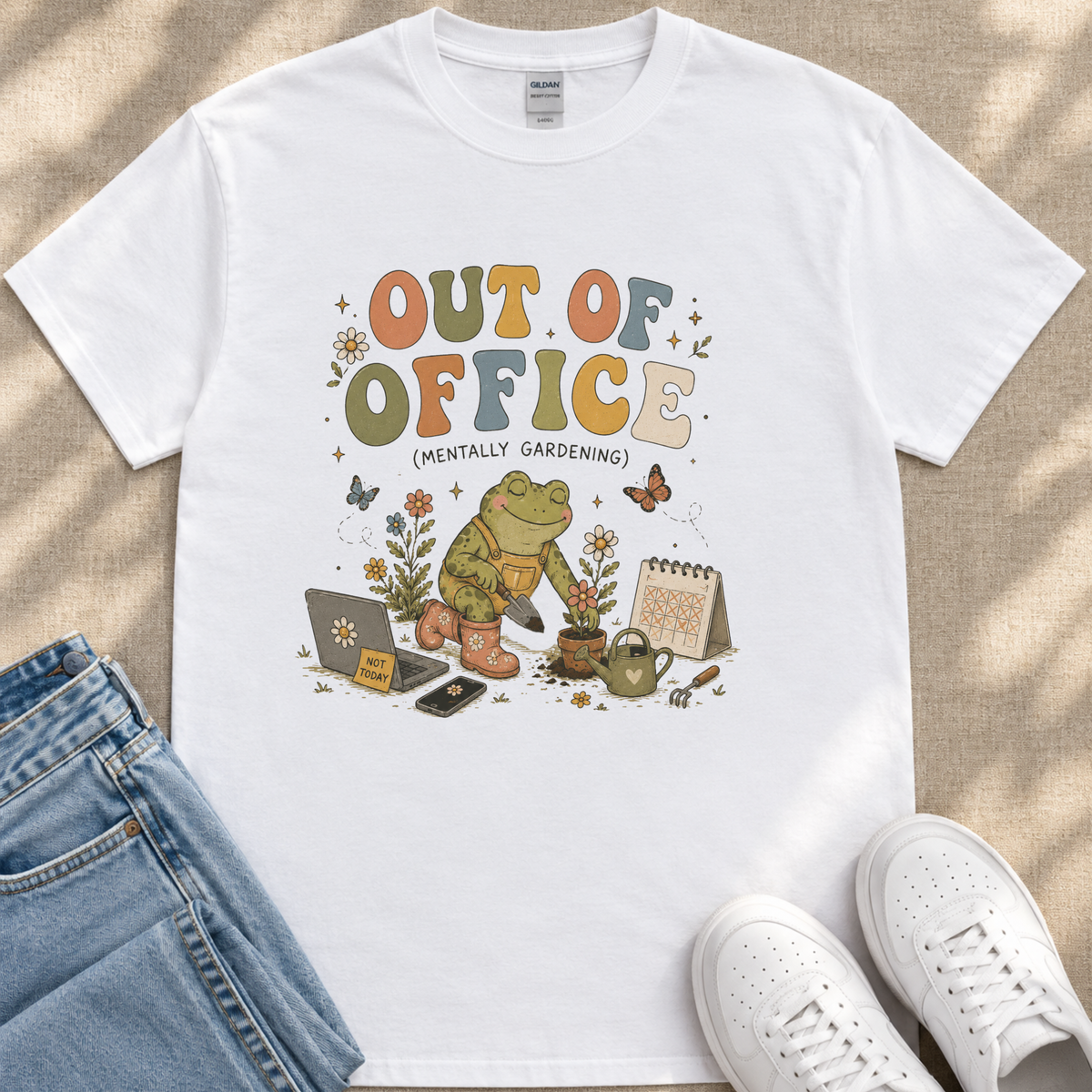 A white t-shirt with a colorful graphic design depicting a frog holding a laptop and surrounded by gardening-related items, along with the text "Out of Office (Mentally Gardening)" and various other elements.
