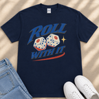 A navy blue t-shirt with a graphic design featuring two dice with smiley faces, along with the text "Roll with it" and a red and blue stripe. The t-shirt is placed on a beige surface, and there are a pair of jeans and white sneakers visible in the image.