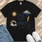 A black t-shirt with a graphic design featuring a sun, clouds, rain, lightning, and a rainbow, along with the text "TAKE IT AS IT COMES" and other weather-related elements.