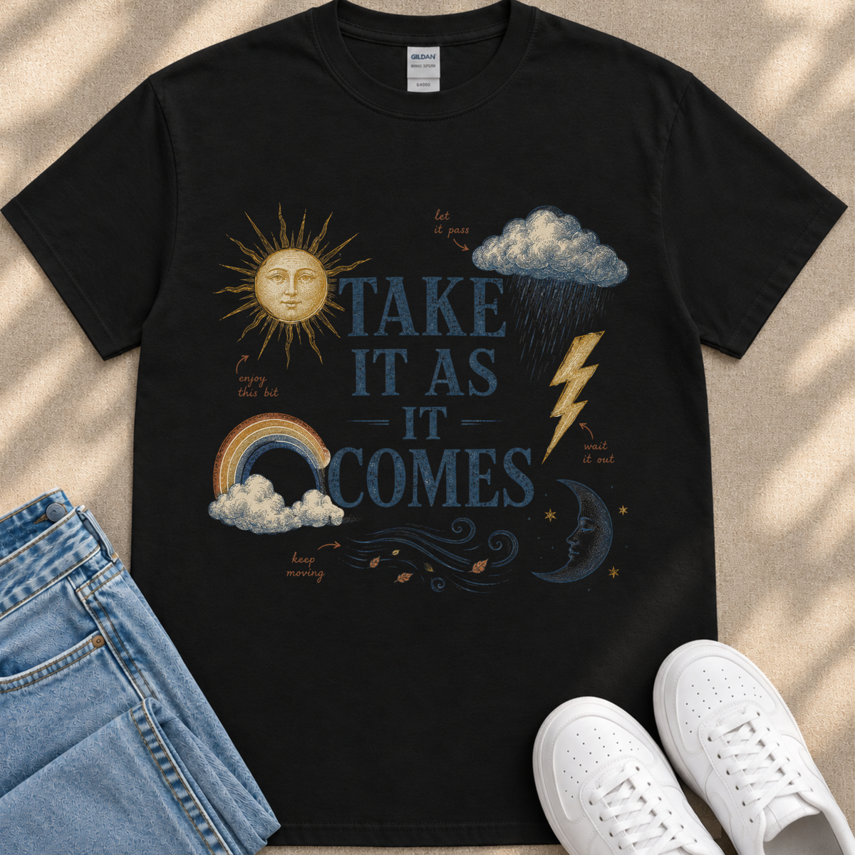 A black t-shirt with a graphic design featuring a sun, clouds, rain, lightning, and a rainbow, along with the text "TAKE IT AS IT COMES" and other weather-related elements.