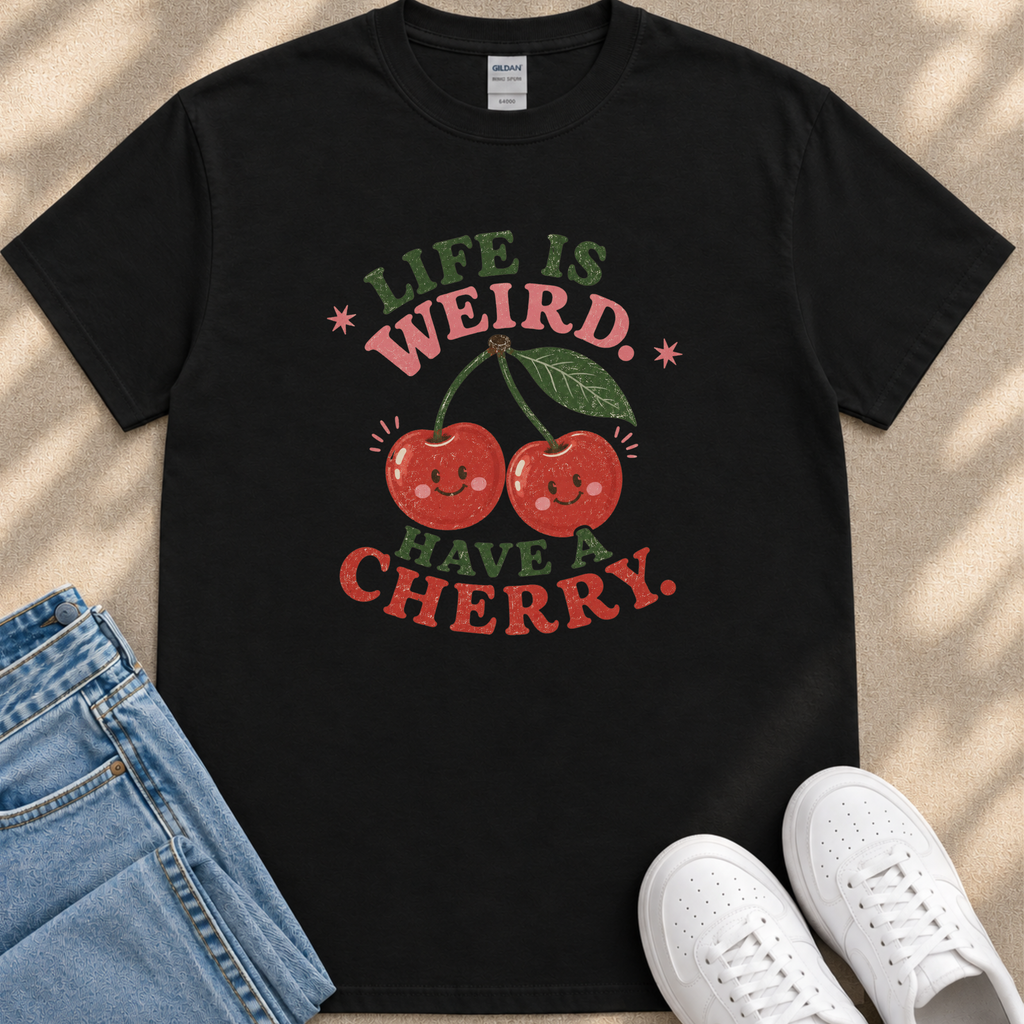a black t-shirt with a graphic design of two cherries and the text "Life is weird. Have a cherry." along with a playful, whimsical font. The t-shirt is placed on a beige surface, and there are a pair of blue jeans and white sneakers visible in the background.
