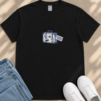 A black t-shirt with a graphic design of a suitcase and a tag that says "Emotional Baggage" is displayed on a beige surface, along with a pair of blue jeans and white sneakers.