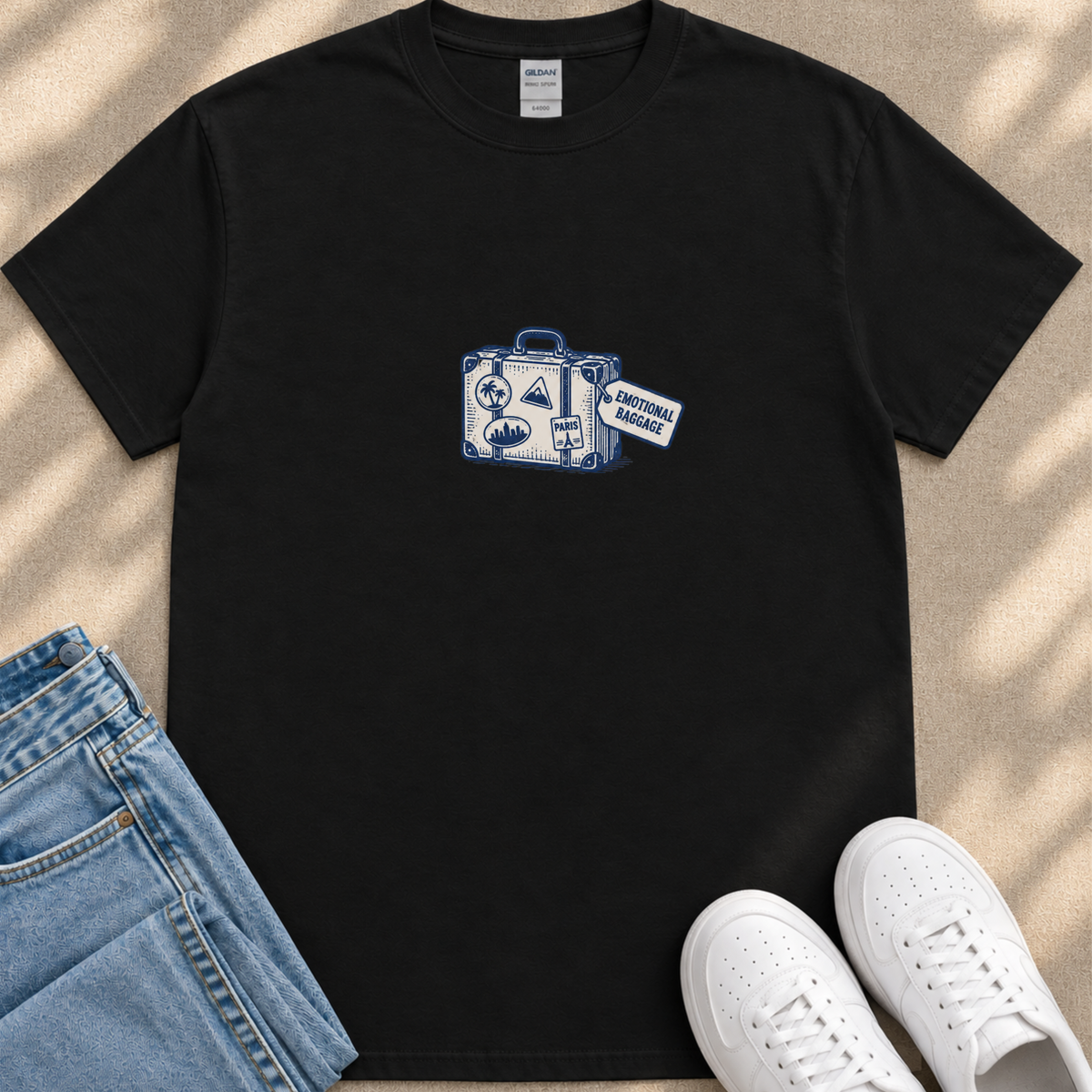 A black t-shirt with a graphic design of a suitcase and a tag that says "Emotional Baggage" is displayed on a beige surface, along with a pair of blue jeans and white sneakers.
