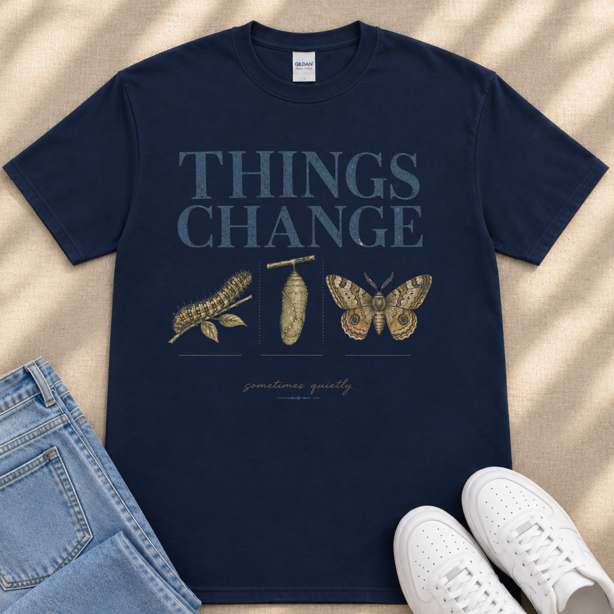 A navy blue t-shirt with the text "THINGS CHANGE" printed on it, along with illustrations of a caterpillar and a butterfly. The t-shirt is laid out on a beige surface, and there are a pair of jeans and white sneakers visible in the image.
