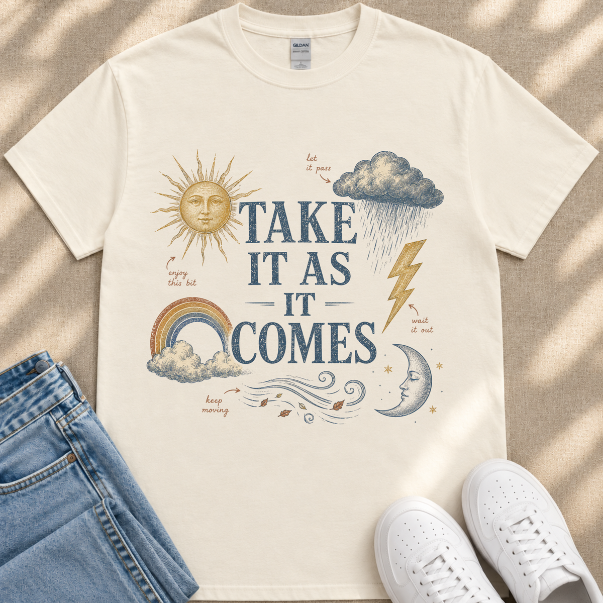 A white t-shirt with a graphic design featuring a sun, clouds, rain, and lightning, along with the text "TAKE IT AS IT COMES" and other weather-related elements.