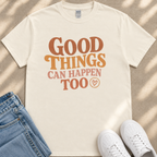 A white t-shirt with the text "GOOD THINGS CAN HAPPEN TO" printed on it, along with a heart symbol. The t-shirt is placed on a beige surface, and there are a pair of blue jeans and white sneakers visible in the image.