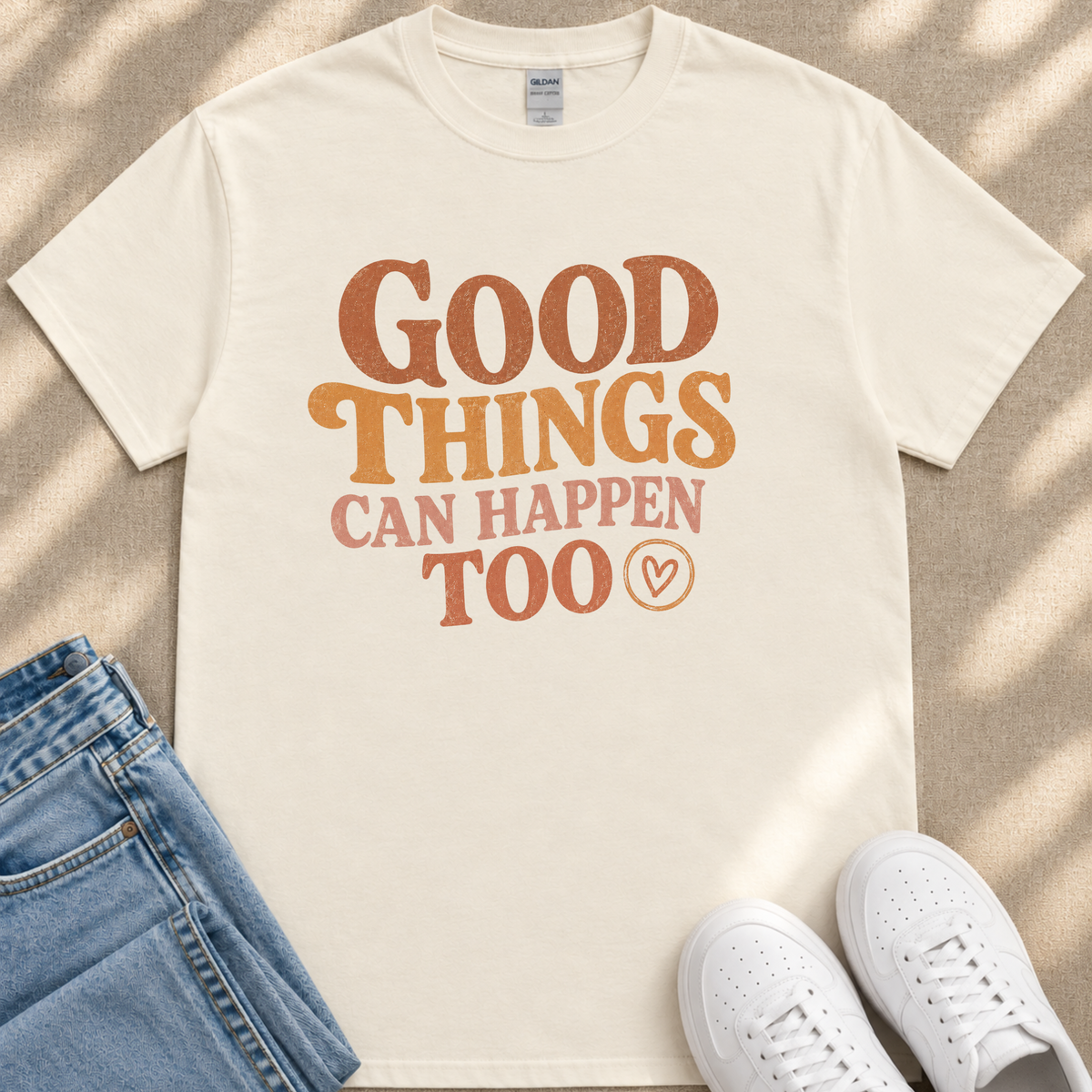 A white t-shirt with the text "GOOD THINGS CAN HAPPEN TO" printed on it, along with a heart symbol. The t-shirt is placed on a beige surface, and there are a pair of blue jeans and white sneakers visible in the image.