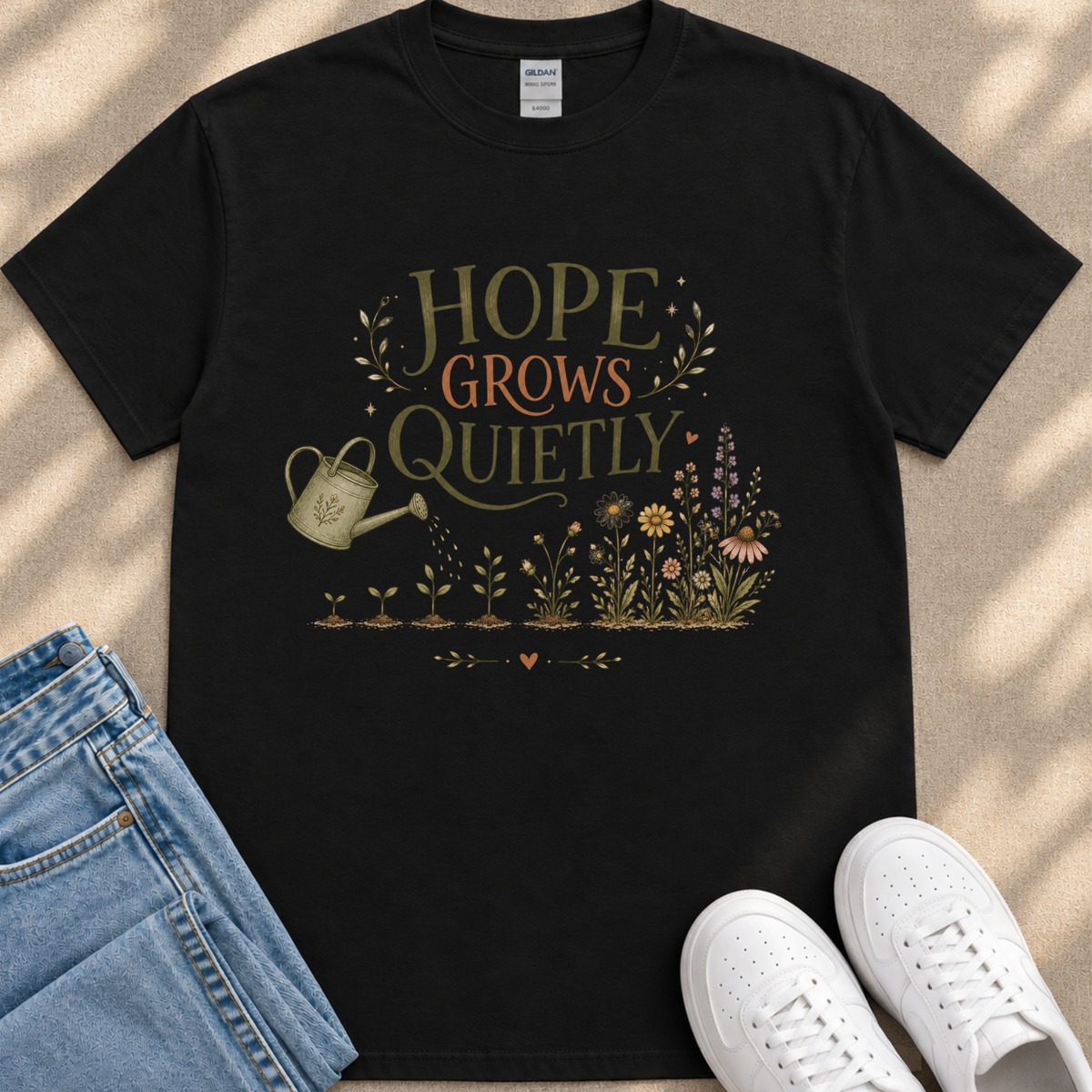 a black t-shirt with a printed design of a watering can surrounded by flowers and plants, along with the text "HOPE GROWS QUICELY". The t-shirt is placed on a beige surface, and there are a pair of blue jeans and white sneakers visible in the image.