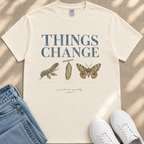A white t-shirt with the text "THINGS CHANGE" printed on it, along with illustrations of a butterfly and a caterpillar. The t-shirt is laid out on a beige surface, and there are a pair of jeans and white sneakers visible in the image.