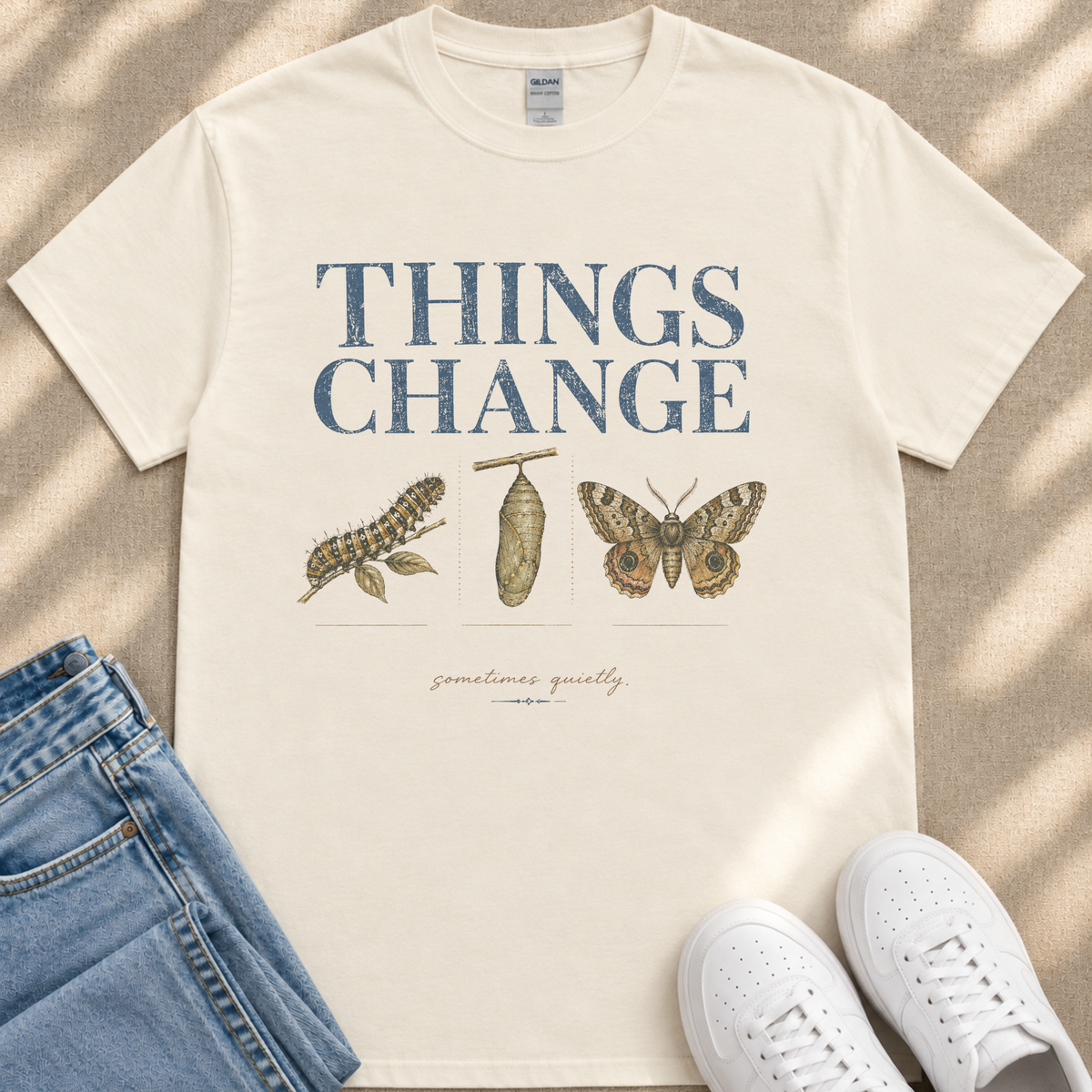 A white t-shirt with the text "THINGS CHANGE" printed on it, along with illustrations of a butterfly and a caterpillar. The t-shirt is laid out on a beige surface, and there are a pair of jeans and white sneakers visible in the image.