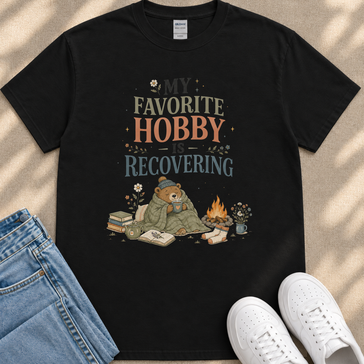 A black t-shirt with a graphic design depicting a bear wrapped in a blanket, sitting by a campfire and surrounded by books and flowers.