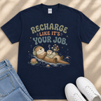 A navy blue t-shirt with a graphic design depicting an otter wearing headphones and floating on a body of water, surrounded by flowers and bubbles. The text on the t-shirt reads "Recharge like it's your job".
