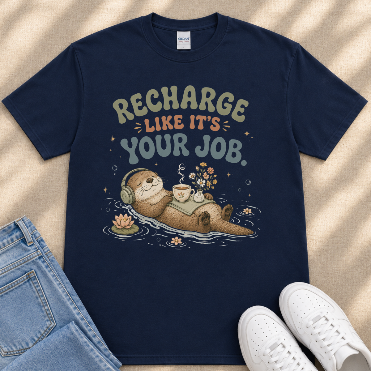 A navy blue t-shirt with a graphic design depicting an otter wearing headphones and floating on a body of water, surrounded by flowers and bubbles. The text on the t-shirt reads "Recharge like it's your job".