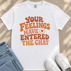 A white t-shirt with a colorful text design that reads "Your Feelings Have Entered The Chat" and a heart symbol. The t-shirt is placed on a beige surface, and there are a pair of blue jeans and white sneakers visible in the image.