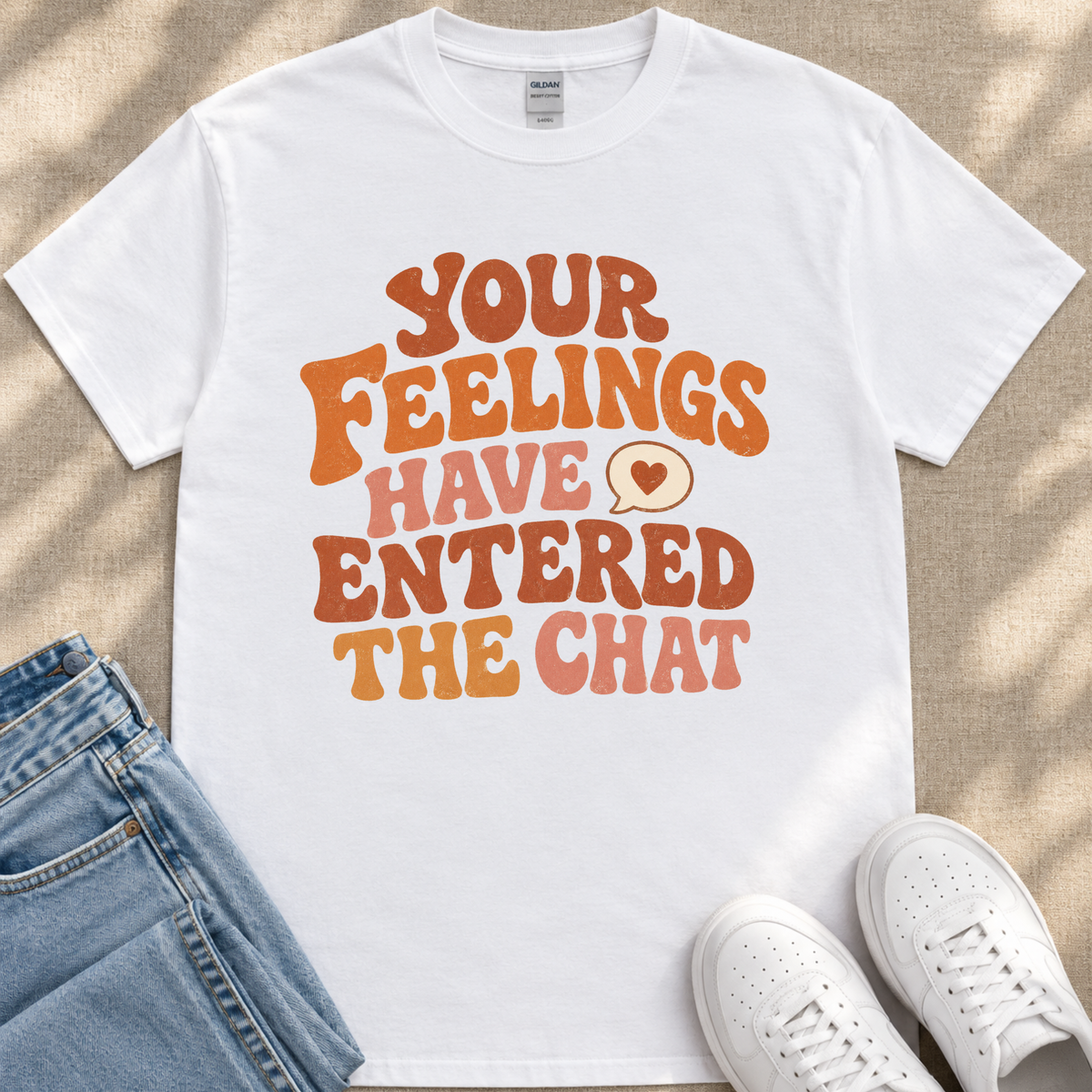A white t-shirt with a colorful text design that reads "Your Feelings Have Entered The Chat" and a heart symbol. The t-shirt is placed on a beige surface, and there are a pair of blue jeans and white sneakers visible in the image.