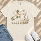a white t-shirt with a printed design of a watering can and flowers, along with the text "HOPE GROWS QUICELY" printed on it. The t-shirt is placed on a beige surface, and there are a pair of blue jeans and white sneakers visible in the image.