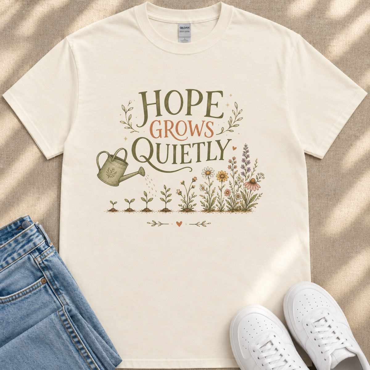 a white t-shirt with a printed design of a watering can and flowers, along with the text "HOPE GROWS QUICELY" printed on it. The t-shirt is placed on a beige surface, and there are a pair of blue jeans and white sneakers visible in the image.