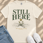 A white t-shirt with the text "STILL HERE" printed on it, along with an illustration of a dandelion seed head. The t-shirt is laid out on a beige surface, and there are a pair of blue jeans and white sneakers visible in the image.