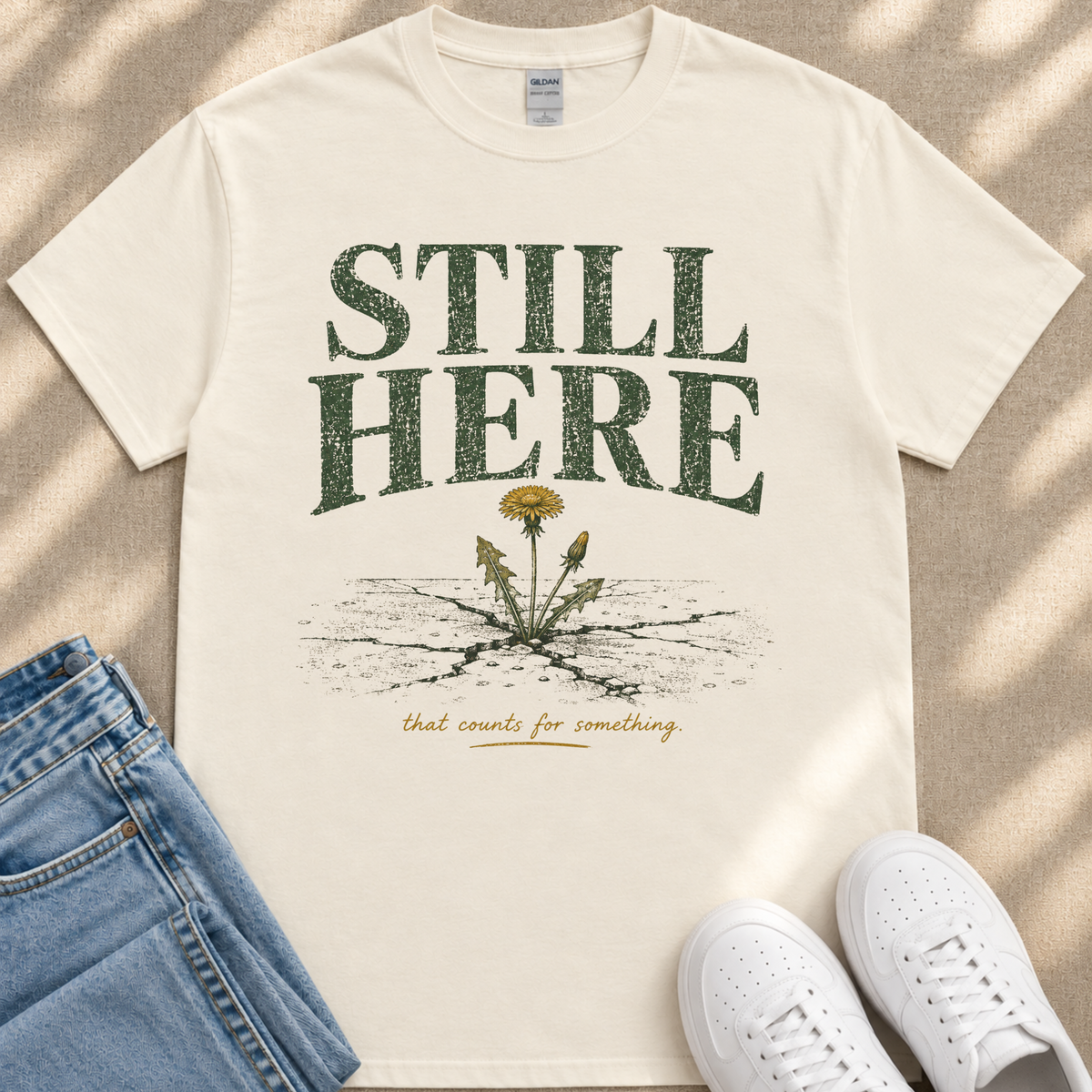 A white t-shirt with the text "STILL HERE" printed on it, along with an illustration of a dandelion seed head. The t-shirt is laid out on a beige surface, and there are a pair of blue jeans and white sneakers visible in the image.