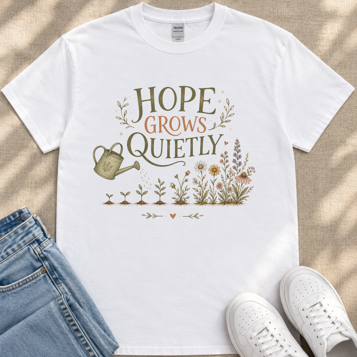 A white t-shirt with a printed design of a watering can and flowers, along with the text "HOPE GROWS QUICELY" printed on it. The t-shirt is placed on a beige surface, and there are a pair of jeans and white sneakers visible in the image.