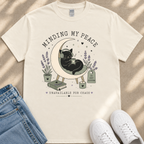 A white t-shirt with a graphic design depicting a black cat sitting on a crescent moon, surrounded by flowers and plants, with the text "MINDING MY PEACE" and "UNAVAILABLE FOR CHAOS" printed on it.
