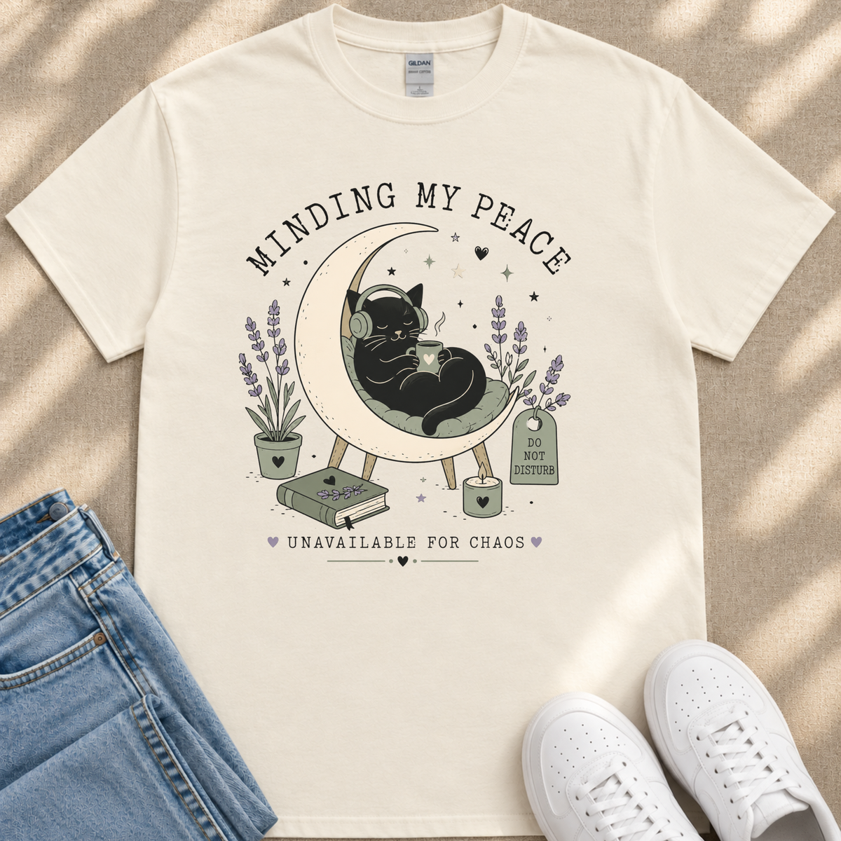 A white t-shirt with a graphic design depicting a black cat sitting on a crescent moon, surrounded by flowers and plants, with the text "MINDING MY PEACE" and "UNAVAILABLE FOR CHAOS" printed on it.
