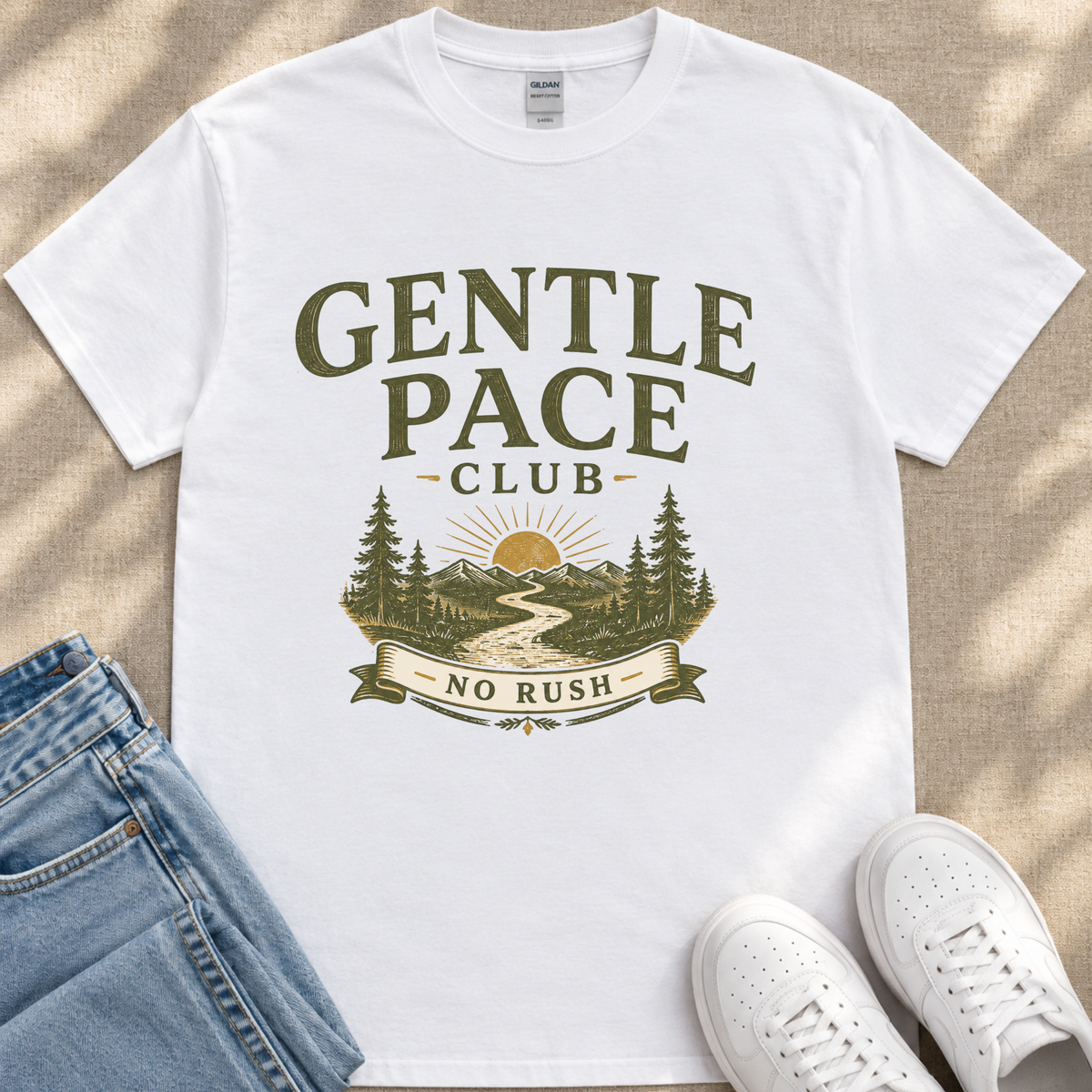 A white t-shirt with a graphic design featuring a mountain landscape, a sun, and the text "GENTLE PACE CLUB NO RUSH" printed on it.