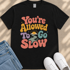 A black t-shirt with a colorful graphic design that says "You're Allowed to Go Slow" in a retro style.