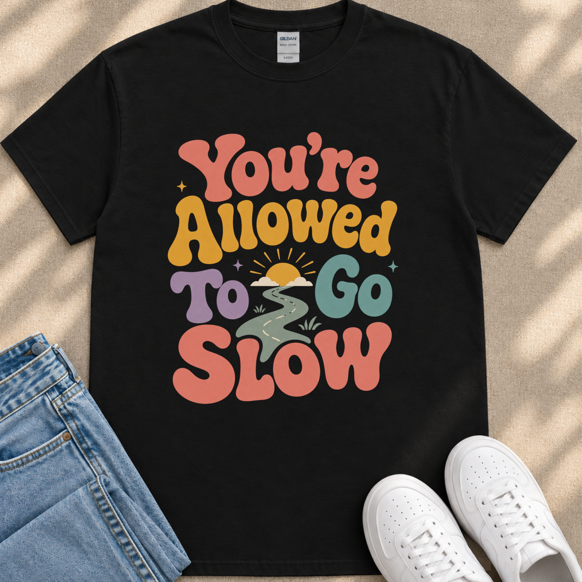 A black t-shirt with a colorful graphic design that says "You're Allowed to Go Slow" in a retro style.