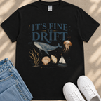 A black t-shirt with a graphic design depicting a whale, jellyfish, and other marine life, along with the text "It's fine to drift". The t-shirt is placed on a beige surface, and there are a pair of jeans and white sneakers visible in the image.