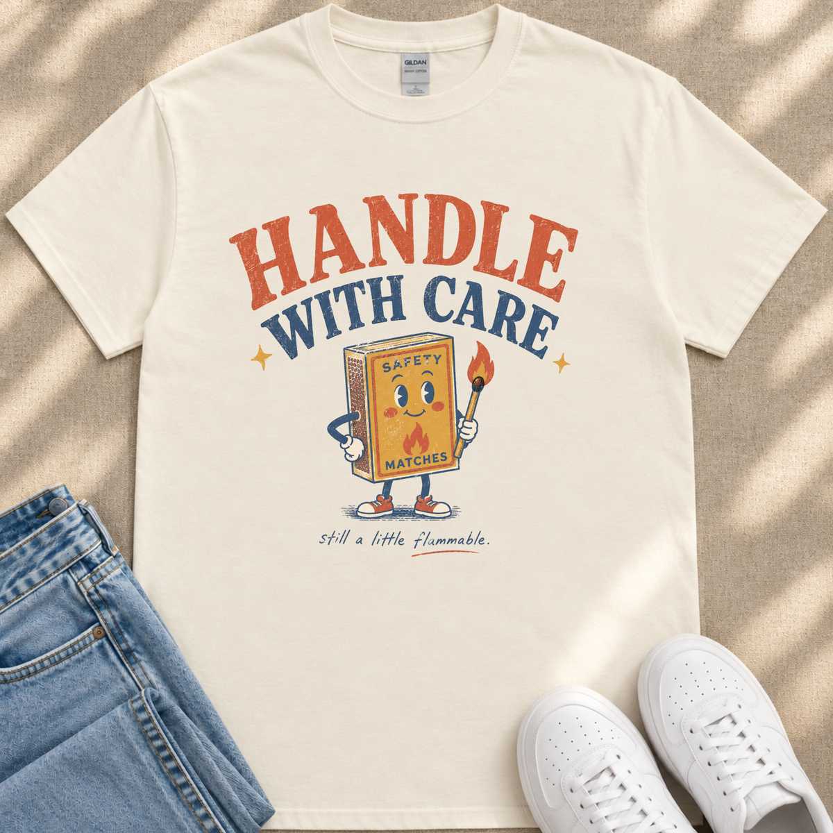 A white t-shirt with a graphic design featuring a cartoon matchbox character holding a lighter and the text "HANDLE WITH CARE" and "SAFETY MATCHES" printed on it.