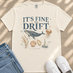 A white t-shirt with a graphic design depicting a whale, jellyfish, and other marine life, along with text that says "It's fine to drift". The t-shirt is laid out on a beige surface, and there are a pair of jeans and white sneakers visible in the image.