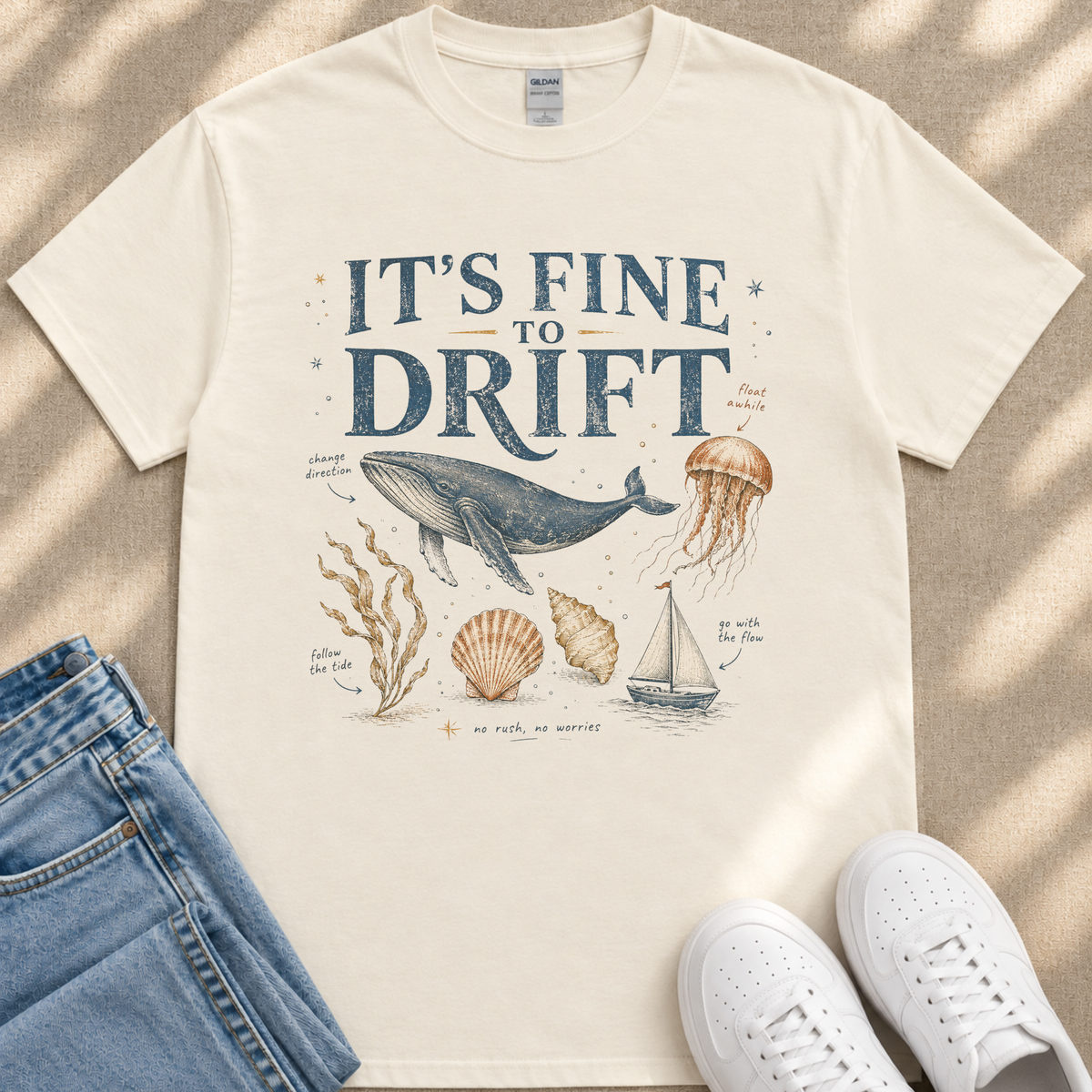A white t-shirt with a graphic design depicting a whale, jellyfish, and other marine life, along with text that says "It's fine to drift". The t-shirt is laid out on a beige surface, and there are a pair of jeans and white sneakers visible in the image.