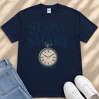 A navy blue t-shirt with the text "SLOW IS SMART" printed on it, along with a clock graphic. The t-shirt is placed on a beige surface, and there are a pair of jeans and white sneakers visible in the image.