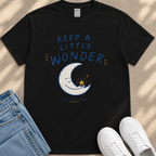 A black t-shirt with a printed design of a crescent moon and stars, along with the text "Keep a little wonder" and a star. The t-shirt is placed next to a pair of jeans and white sneakers.