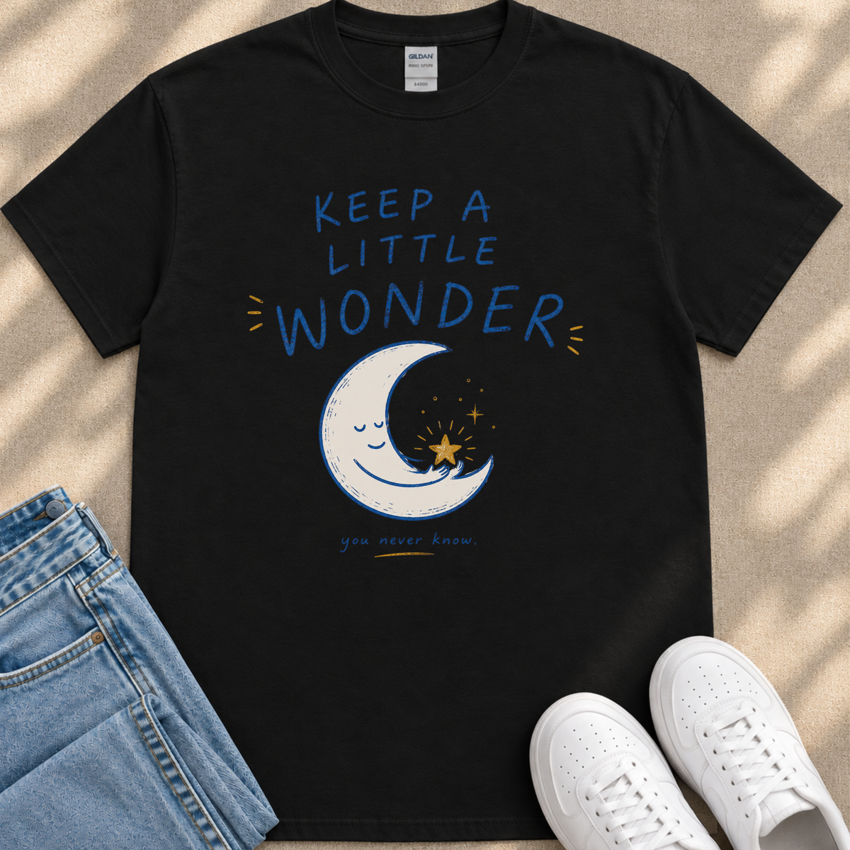 A black t-shirt with a printed design of a crescent moon and stars, along with the text "Keep a little wonder" and a star. The t-shirt is placed next to a pair of jeans and white sneakers.