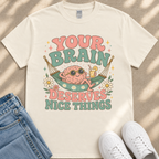 A white t-shirt with a colorful graphic design featuring a brain character in a hammock, surrounded by flowers and text that reads "Your brain deserves nice things".