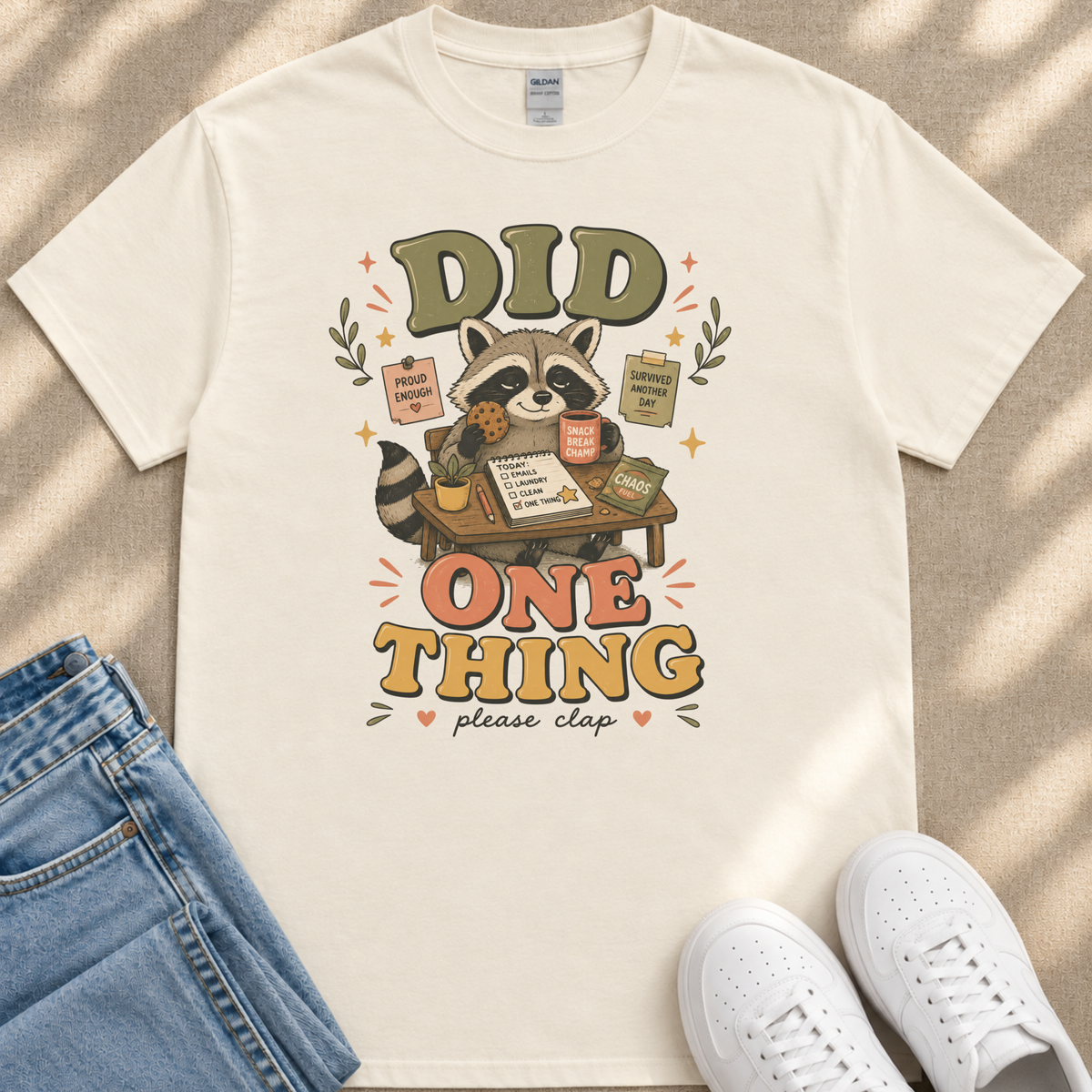 A white t-shirt with a colorful graphic design featuring a raccoon holding a sign that says "Did One Thing" and surrounded by various objects like a book, a cup, and a plant.