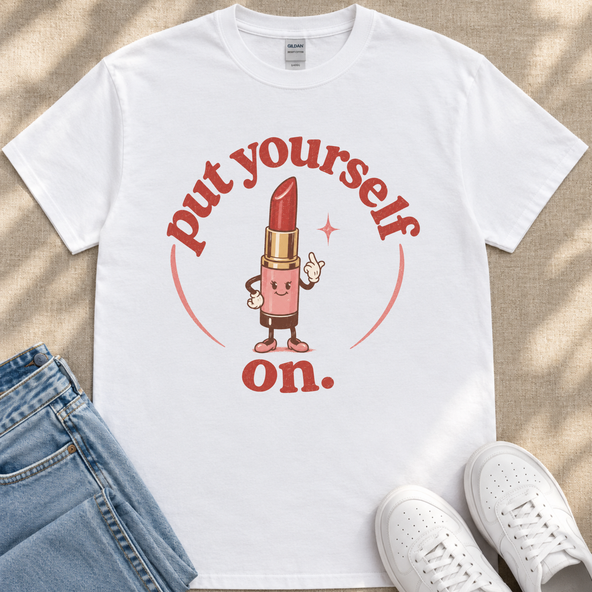 A white t-shirt with a graphic design featuring a lipstick character and the text "put yourself on."
