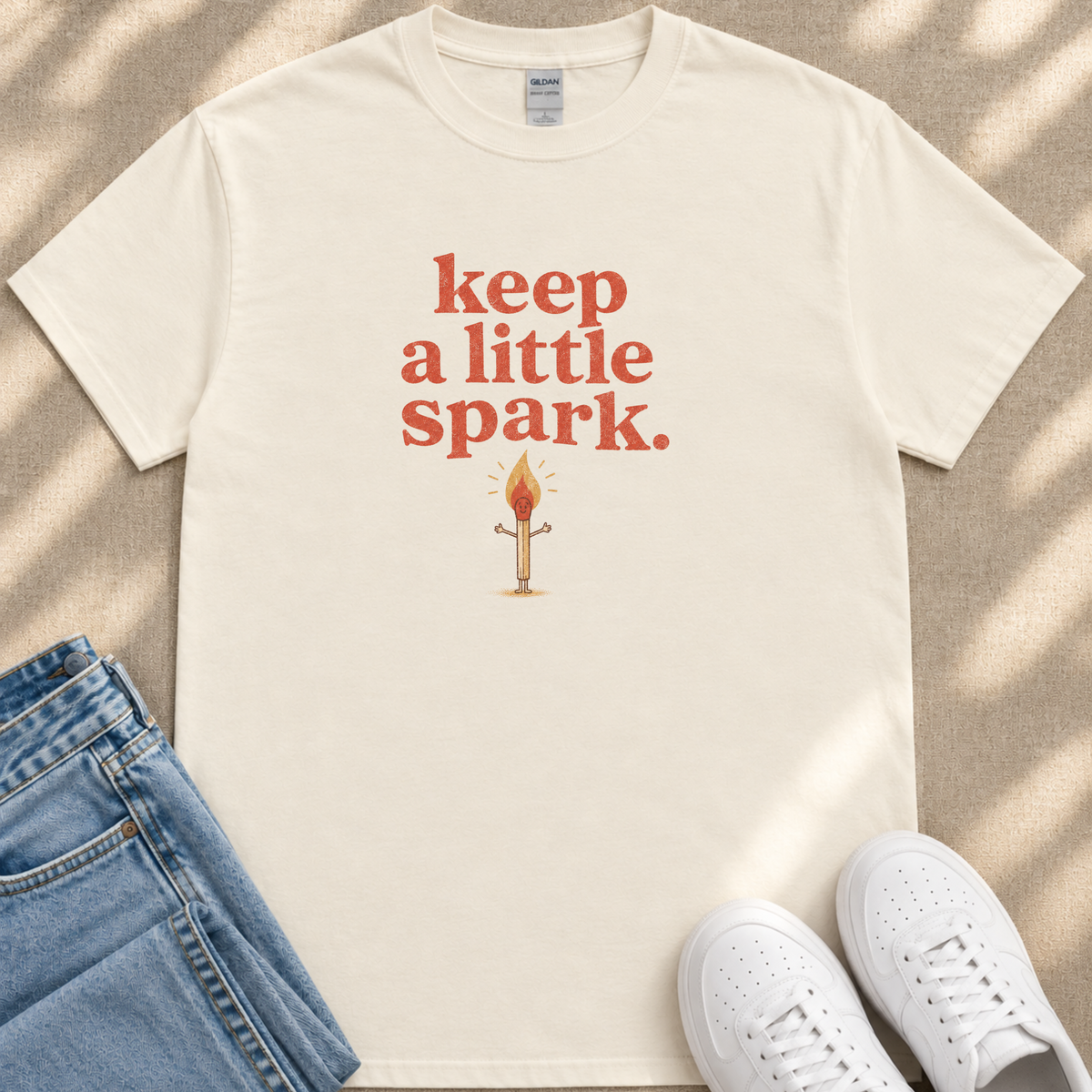 A white t-shirt with the text "keep a little spark" printed on it, along with an illustration of a lit match. The t-shirt is placed next to a pair of blue jeans and white sneakers.