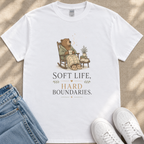 A white t-shirt with a graphic design depicting a bear sitting in a rocking chair, holding a cup and surrounded by potted plants. The text below the image reads "Soft Life, Hard Boundaries."