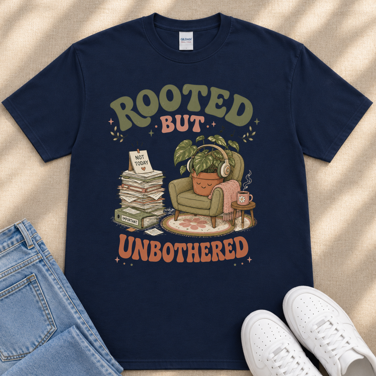 A navy blue t-shirt with a graphic design depicting a potted plant, a stack of books, and a person wearing headphones, along with the text "ROOTED BUT UNBOTHERED" printed on it.