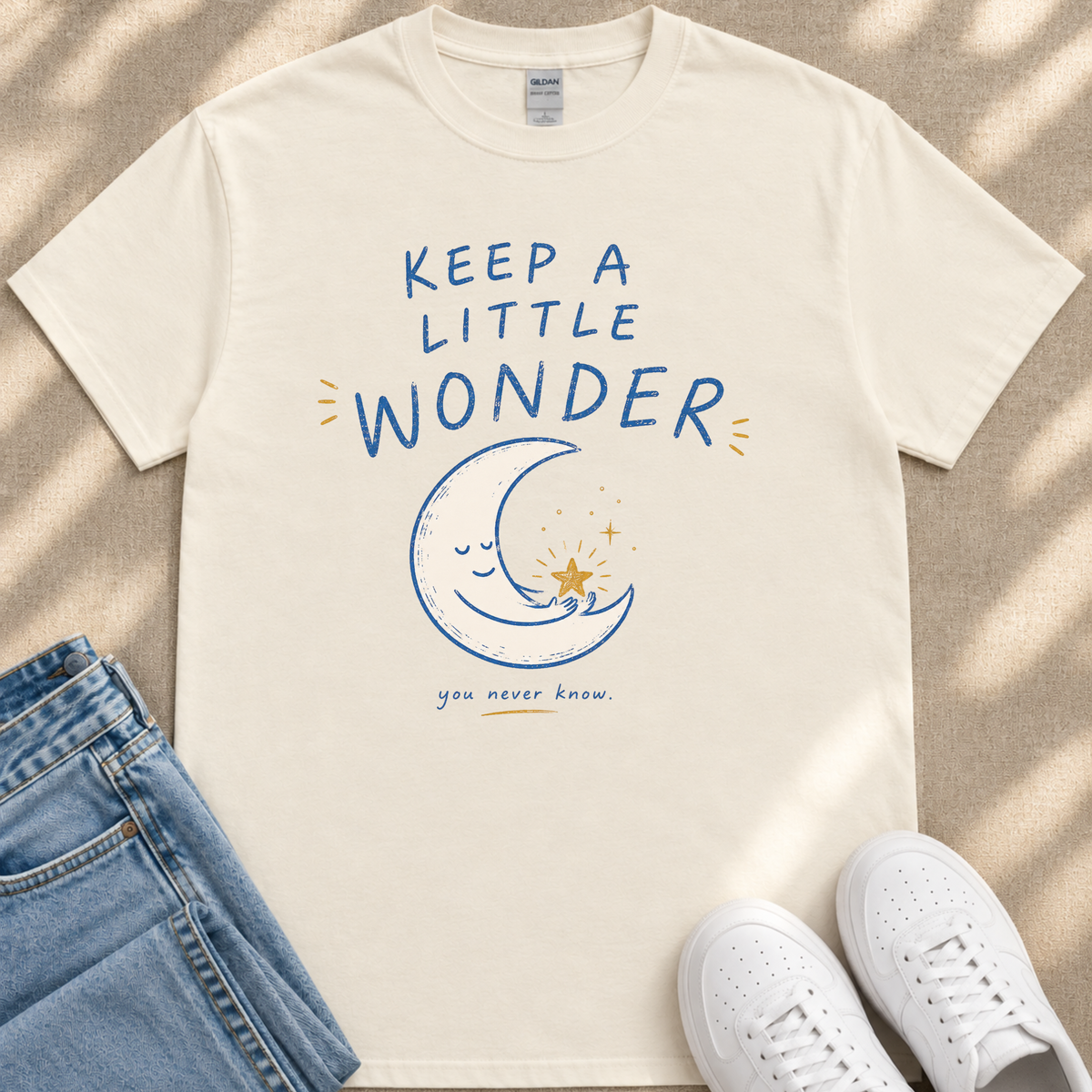 A white t-shirt with a blue moon and star graphic, along with the text "KEEP A LITTLE WONDER YOU NEVER KNOW" printed on it. The t-shirt is placed next to a pair of blue jeans and white sneakers.