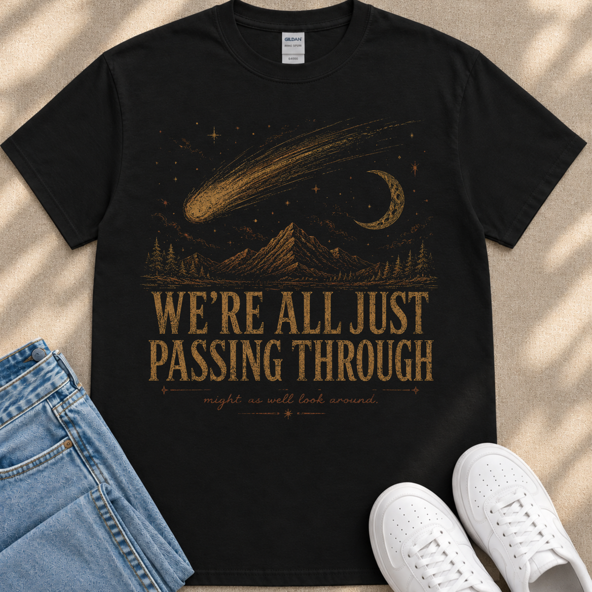 A black t-shirt with a graphic design depicting a shooting star, a crescent moon, and mountains, along with the text "WE'RE ALL JUST PASSING THROUGH" and "MIGHT AS WELL LOOK AROUND." The t-shirt is placed on a beige surface, and there are a pair of blue jeans and white sneakers visible in the image.