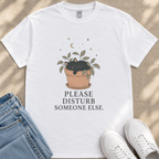 A white t-shirt with a graphic design depicting a cat sleeping in a pot with plants and stars above it, along with the text "PLEASE DISTURB SOMEONE ELSE."