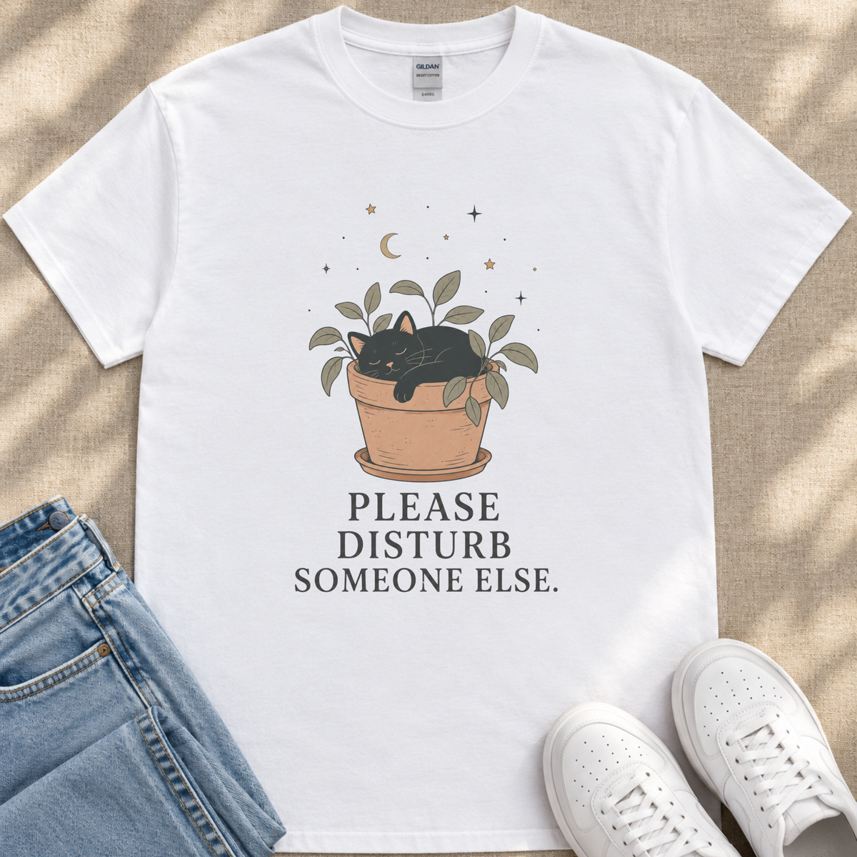 A white t-shirt with a graphic design depicting a cat sleeping in a pot with plants and stars above it, along with the text "PLEASE DISTURB SOMEONE ELSE."